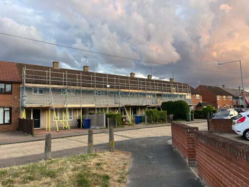 Row of houses with scaffolding, surrounded by green hedges and a cloudy sky.