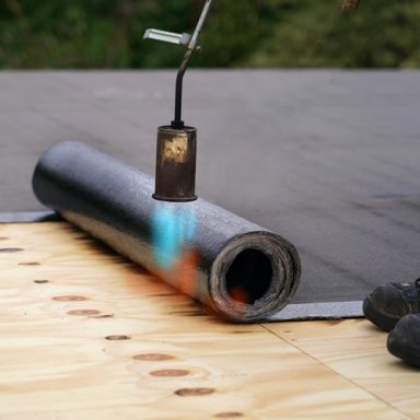 A propane torch heating a roll of roofing material on a wooden surface.