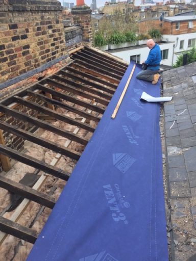 A person measuring on a roof with exposed wooden beams and a blue tarp.