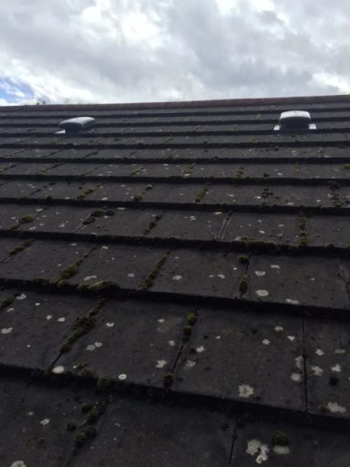 Close-up of a dark roof with moss and two ventilation vents under a cloudy sky.